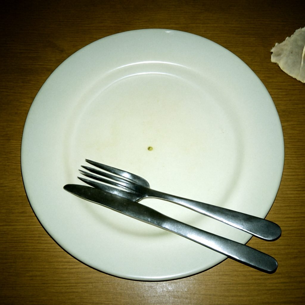A harsh flash photo shot straight down at a wiped-clean white dinner plate with a single crumb left on it, knife and fork laid together across the rim in the finished-eating position, on a scuffed wood table with a crumpled napkin to one side